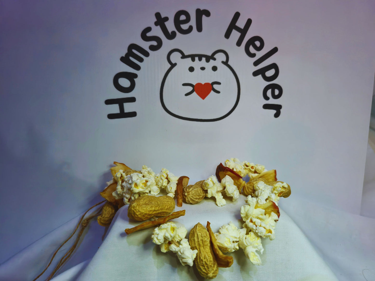 A hamster chewy garland containing popcorn, monkey nuts and apple on a string in front of the Hamster Helper logo.