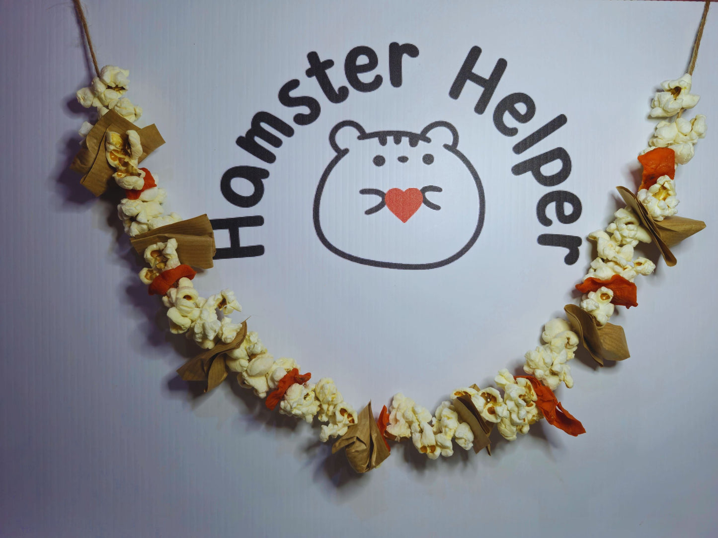 A hamster garland with popcorn, hazelnuts and dried carrots on a string hanging beneath the hamster helper logo.