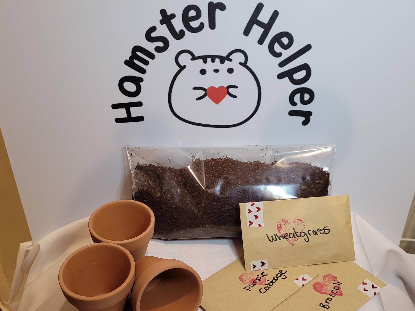 A kit for growing microgreens containing three terracotta pots, three packets of seeds and a bag of hamster safe soil arranged in front of the Hamster Helper logo