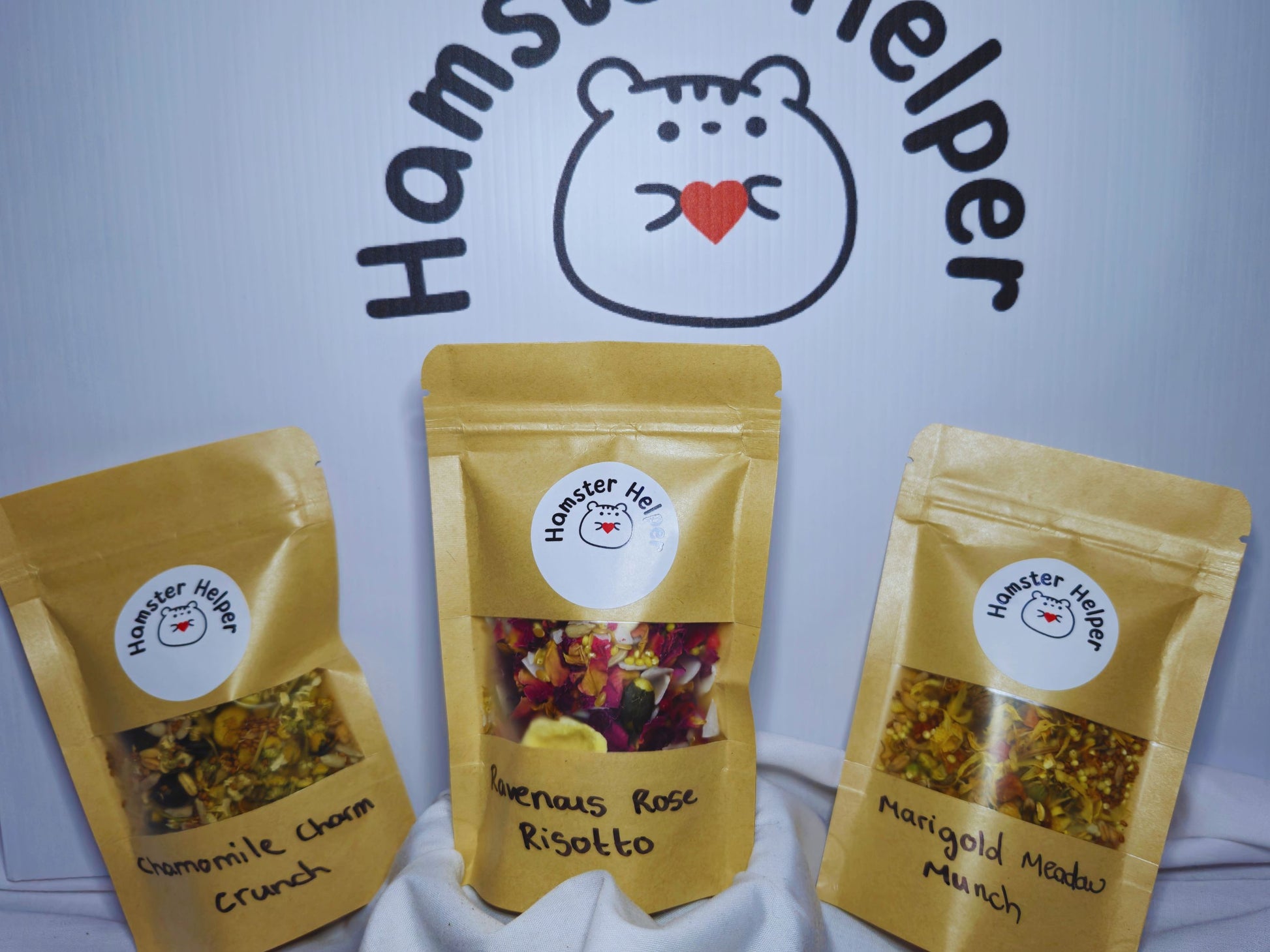 Three mini hamster treat mixes arranged in front of the hamster helper logo. Each treat mix is in a pouch with a Hamster Helper sticker on it. The treat mixes are chamomile charm crunch, ravenous rose risotto and marigold meadow munch.