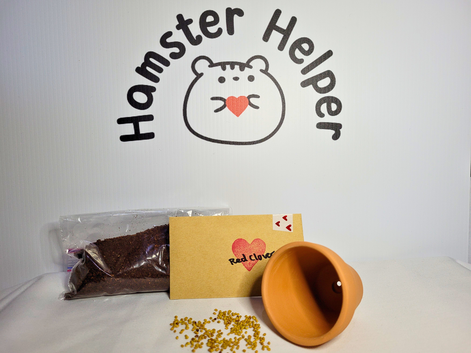 A hamster safe red clover microgreen kit containing a small bag of soil, pack of red clover seeds and a terracotta pot displayed in front of the Hamster Helper logo