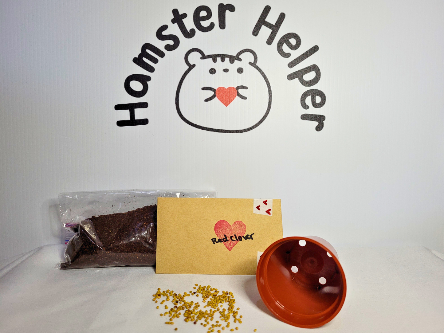 A kit for growing hamster safe red clover microgreens containing hamster safe soil, a plastic plant pot and some red clover seeds in front of the Hamster Helper logo